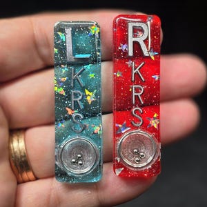 May include: Two rectangular resin pieces, one teal and one red, each featuring embedded silver letters spelling LKRS vertically. Both pieces contain holographic star-shaped glitter and a circular metal bowl at the base with three small silver beads.
