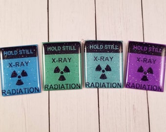 May include: Four rectangular, glittery magnets in blue, green, teal, and purple. Each magnet has the words "HOLD STILL", "X-RAY", and "RADIATION" with a radiation symbol. The magnets are on a white wooden surface.