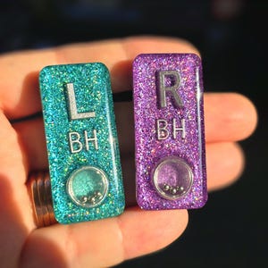 May include: Two rectangular, glittery resin keychains. One is teal with the letter "L" and the letters "BH", the other is purple with the letter "R" and the letters "BH". Each has a small, clear, circular element with tiny silver beads.