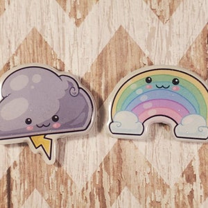 May include: Two resin charms, one shaped like a purple cloud with a lightning bolt and a smiling face, the other shaped like a rainbow with a smiling face and white clouds on either side.