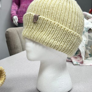 May include: A light yellow ribbed knit beanie hat with a folded brim. The hat is displayed on a white mannequin head. A small brown leather tag with the letters "AV" is attached to the brim.