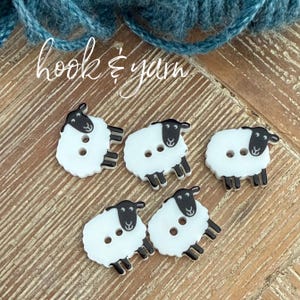 May include: Five white and black sheep-shaped buttons, each featuring two holes for attachment. The buttons are arranged in a group, displaying a detailed black head and legs against a white body. The text hook & yarn is visible above.