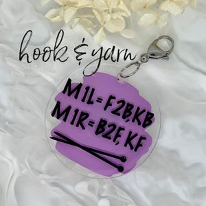 May include: Purple acrylic keychain with a black hook and a silver split ring. The keychain has a purple background with black text that reads "MIL=F2BKB MIR=B2F,KF" and a pair of black knitting needles.