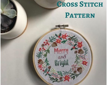May include: A cross stitch pattern of a wreath with red berries, green leaves, and brown pinecones. The wreath surrounds the words "Merry and Bright".