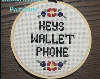 May include: A white cross stitch embroidery hoop with a wooden frame. The embroidery features black text that reads "KEYS WALLET PHONE" with red, yellow, and blue flowers at the top and bottom.