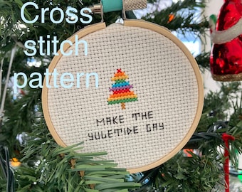 May include: A rainbow-coloured Christmas tree cross stitch ornament with the text "Make the Yuletide Gay" hanging on a green Christmas tree.