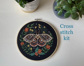 May include: A cross stitch kit featuring a moth with brown, black, and white wings surrounded by a floral wreath with red, yellow, and green flowers. The kit is on a black background.