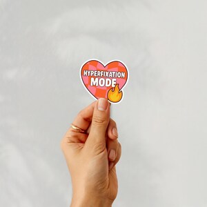 May include: A heart-shaped sticker with the words "HYPERFIXATION MODE" in a bubble font. The sticker has a pink and orange checkered pattern and a small flame graphic. The sticker is held up by a hand.