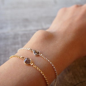 May include: Two delicate bracelets featuring small, teardrop-shaped gemstones. The bracelet on the left is gold with a round gemstone, while the bracelet on the right is silver with a teardrop-shaped gemstone.