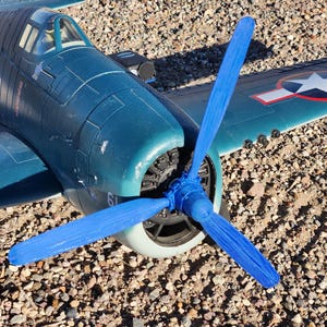 May include: A teal-colored model airplane with a blue propeller. The plane has a white star and red circle emblem on the wing. The model is sitting on a bed of small rocks.