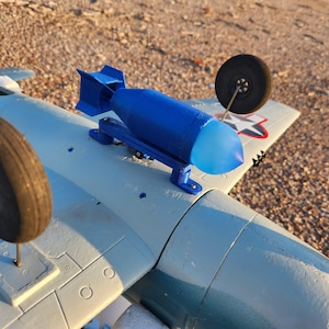 Puede incluir: Un modelo de avión azul claro con una bomba azul adjunta. El avión tiene ruedas negras e insignias de estrella roja, blanca y azul en las alas. El modelo está sobre un fondo de pequeños guijarros.