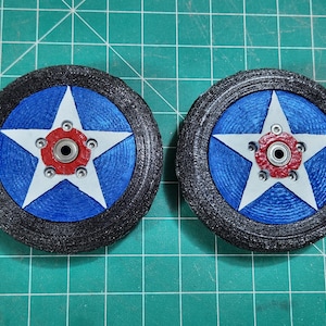 Peut inclure: Deux roues de modèle avec un centre bleu orné d'une étoile blanche, un moyeu rouge et un pneu noir. Les roues présentent un aspect texturé et détaillé, sur un fond de grille verte.