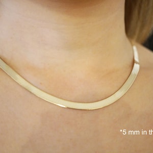 May include: A gold-colored, flat herringbone chain necklace, approximately 5 mm wide, resting on a person's neck. The necklace has a smooth, reflective surface, and the image is well-lit, highlighting the jewelry's design and color.