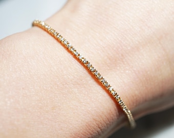 May include: A delicate gold bracelet adorned with small, sparkling gemstones. The bracelet is thin and elegant, resting gently on a wrist. The gemstones are evenly spaced along the gold band, creating a continuous line of sparkle.