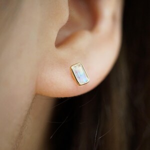 Puede incluir: Primer plano de un pendiente de botón rectangular dorado con una piedra preciosa rectangular iridiscente. El pendiente está engastado en un bisel dorado y se lleva en una oreja.