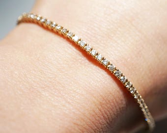 May include: A gold bracelet with a row of sparkling diamonds. The bracelet is set on a gold band and is worn on a wrist. The diamonds are small and evenly spaced, creating a continuous line of brilliance.