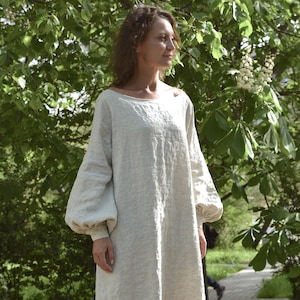 May include: A light beige linen dress with long, voluminous sleeves. The dress has a boat neckline and a floor-length hem. The model is standing on a wooden surface in a natural outdoor setting, surrounded by green foliage.