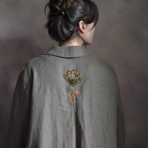 May include: A long, olive-green coat with a decorative floral design on the back. The flower is embellished with gold and green beads. The coat has a classic collar and a relaxed fit, suitable for layering.