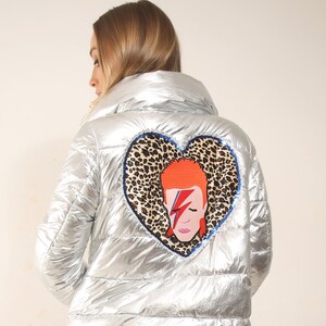 May include: A silver puffer jacket with a heart-shaped patch featuring a portrait of David Bowie on a leopard print background. The jacket is worn over a leopard print skirt and black fishnet tights.