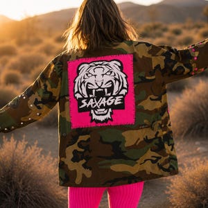 May include: A camouflage jacket featuring a large, square, hot pink patch on the back. The patch displays a black and white graphic of a snarling tiger face with the word SAVAGE printed in bold, black, capital letters across the tiger's mouth.