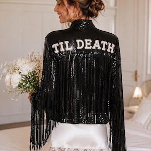 Puede incluir: Una chaqueta negra de lentejuelas con flecos largos y el texto TIL DEATH bordado en letras blancas en la parte superior de la espalda. La chaqueta se lleva sobre un vestido lencero de satén blanco con un delicado borde de encaje en el dobladillo.