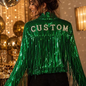 May include: A bright green sequined jacket featuring long fringe detailing across the back and sleeves. The word CUSTOM is spelled out in white pearl-like letters across the upper back. The jacket has a high collar and a full-length sequin finish.