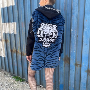 May include: A black hooded jacket with a blue and black tiger-striped faux fur vest. The vest features a white tiger face graphic and the word "SAVAGE" in white. The person is wearing black boots and fishnet stockings.