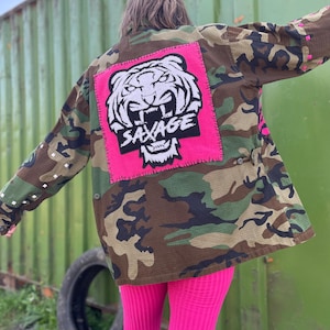 May include: A camouflage jacket with a large pink patch featuring a white tiger illustration and the word "SAVAGE" in black. The jacket has a button-up front and is paired with bright pink leggings. The background is a green metal structure.