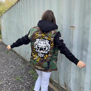 May include: A camouflage jacket with black sleeves and a black hood. The back of the jacket features a white tiger head with the word "SAVAGE" and yellow stripes. The person is wearing white pants and black boots.
