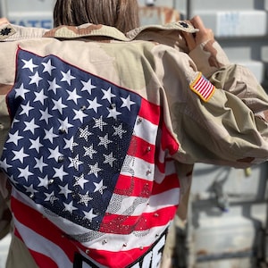 May include: Camouflage jacket with an American flag design on the back. The flag features white stars on a blue field, red and white stripes, and the word "LOVE" in sequins. An American flag patch is on the sleeve.