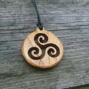 Puede incluir: Un collar con un colgante de madera con un diseño de nudo celta. El colgante es redondo y tiene una veta de madera marrón oscura. El collar está hecho con un cordón negro.