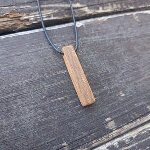 May include: A wooden pendant necklace with a rectangular, light brown pendant hanging from a dark grey cord. The pendant has a natural wood grain texture. The necklace is shown on a weathered wooden surface.