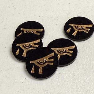 May include: A collection of black, circular game tokens featuring a gold-colored Eye of Horus symbol. The tokens are arranged on a light-colored surface, with the gold design contrasting against the black background. These tokens are likely for a board game.