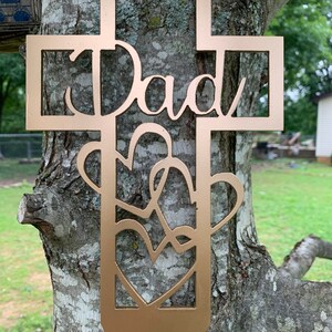 以下が含まれることがあります： 中央に「Dad」という文字がくり抜かれた金色の金属製の十字架。十字架は、絡み合ったハートのデザインで飾られています。