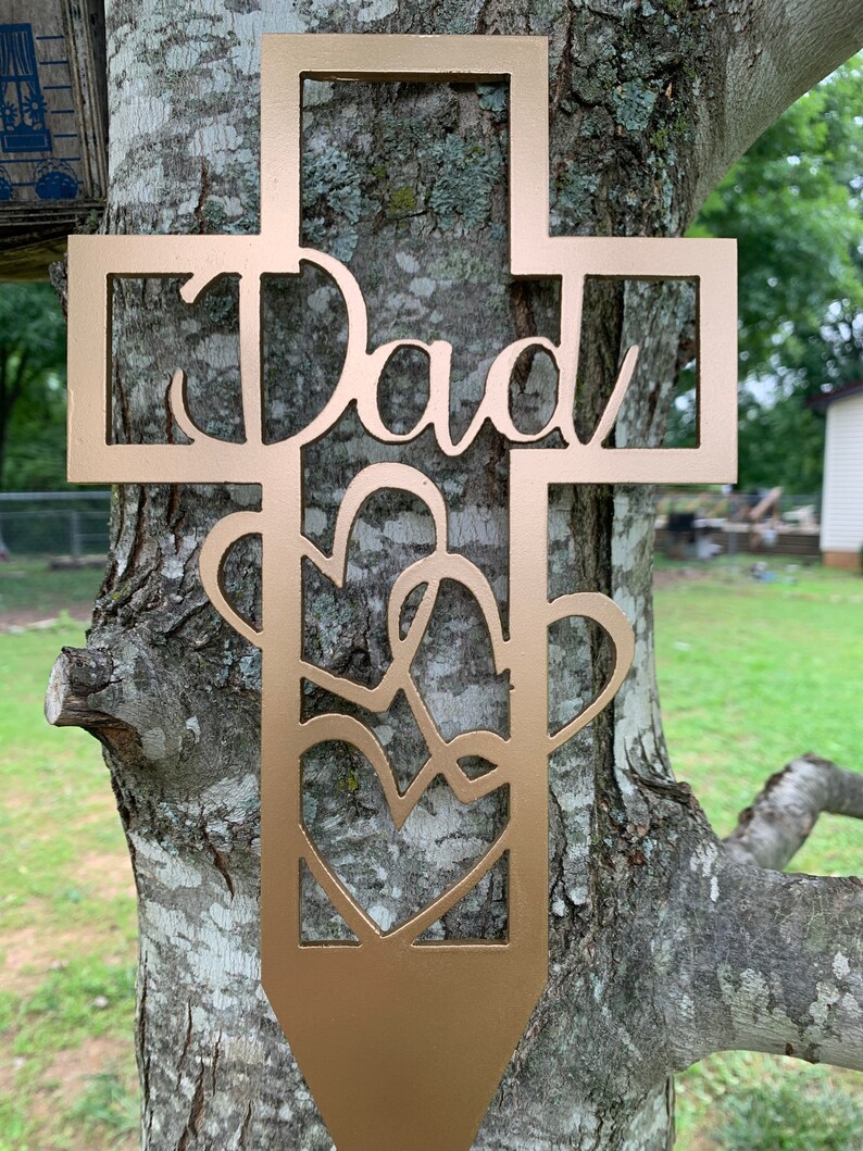 以下が含まれることがあります： 中央に「Dad」という文字がくり抜かれた金色の金属製の十字架。十字架は、絡み合ったハートのデザインで飾られています。