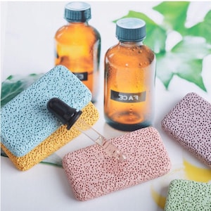 Op de afbeelding: Een assortiment gekleurde puimstenen in blauw, geel, roze en groen, samen met twee bruine glazen flessen met donkere doppen. Een glazen pipet ligt op de stenen, wat wijst op huidverzorgings- of schoonheidsproducten.