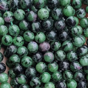 Puede incluir: Primer plano de una colección de cuentas redondas de piedras preciosas. Las cuentas son una mezcla de colores verde, negro y rosa-púrpura. Las cuentas son lisas y pulidas, con un aspecto natural y orgánico. Las cuentas se utilizan probablemente para la fabricación de joyas.