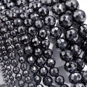 May include: Close-up of faceted, dark gray hematite beads. The beads have a metallic sheen and are strung together, showcasing their geometric, multi-faceted surfaces. The beads vary in size, creating a textured, layered effect.