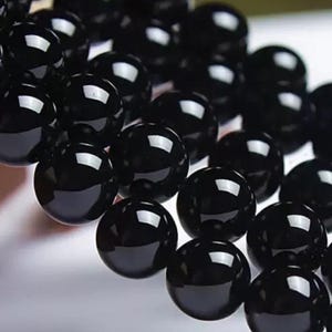 Può includere: Primo piano di una collezione di perline sferiche nere e lucide. Le perline sono fittamente raggruppate, riflettendo la luce e creando una superficie lucida. Sembrano essere infilate, suggerendo una collana o un braccialetto.