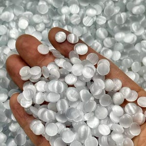Può includere: Primo piano di una mano che tiene piccole pietre rotonde di selenite bianca. Le pietre sono lisce e traslucide, con una sottile lucentezza. Lo sfondo è pieno di altre pietre simili. Queste pietre sono spesso utilizzate per la guarigione e la meditazione.