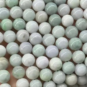 Puede incluir: Primer plano de una colección de cuentas redondas y pulidas. Las cuentas son una mezcla de blanco, verde pálido y gris, con un acabado brillante. Las cuentas probablemente están hechas de jade o una piedra similar, y están ensartadas.