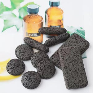 Puede incluir: Una variedad de piedras pómez de color gris oscuro en varias formas, incluyendo redondas, ovaladas y rectangulares. Dos botellas de color ámbar con tapas azules en el fondo. Una botella tiene la palabra "FACE" impresa.