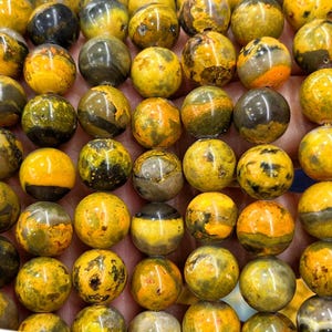 Pode incluir: Close-up de uma coleção de contas redondas e polidas de pedras preciosas. As contas são principalmente amarelas com veios pretos, castanhos e laranja. As contas estão dispostas próximas umas das outras, preenchendo a moldura.