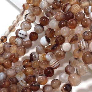 Puede incluir: Una colección de cuentas de piedras preciosas marrones y beige. Las cuentas varían en color, desde beige claro hasta marrón oscuro, algunas con patrones de rayas. Las cuentas son redondas y parecen estar ensartadas, ideales para la creación de joyas.