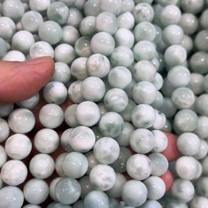以下が含まれることがあります： 丸い薄緑色のビーズの山をクローズアップ。ビーズは白と濃い緑色の渦巻き模様で大理石のような外観です。ビーズは滑らかで光沢があり、研磨された仕上がりを示唆しています。ジュエリー作りや工芸品に適しています。