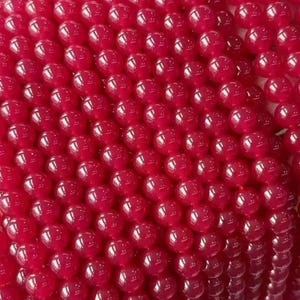 Op de afbeelding: Close-up van een verzameling levendige rode, ronde kralen. De kralen zijn dicht op elkaar gepakt, waardoor een textuur ontstaat. De glanzende afwerking van de kralen reflecteert het licht, wat diepte aan de afbeelding toevoegt. Ideaal voor het maken van sieraden.