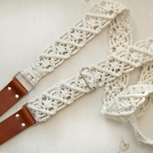 Może przedstawiać: Kremowy pasek do gitary z makramy z brązowymi skórzanymi końcówkami i srebrnym osprzętem. Pasek wykonany jest z tkanej bawełnianej liny i ma dekoracyjny wzór. Stylowy dodatek dla muzyków.