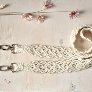 May include: A cream-colored macrame phone strap featuring an intricate woven diamond pattern. The strap is attached to a white phone case with two silver-toned metal lobster claw clasps, allowing for a secure connection to the mobile device.