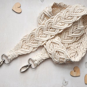 Puede incluir: Una correa de macramé de algodón blanco con cierres de metal plateado. La correa está tejida en un patrón de diamante.