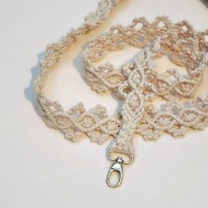 May include: A white crocheted strap with a gold metal clasp. The strap is made of a thick, textured cord and has a delicate, intricate design.
